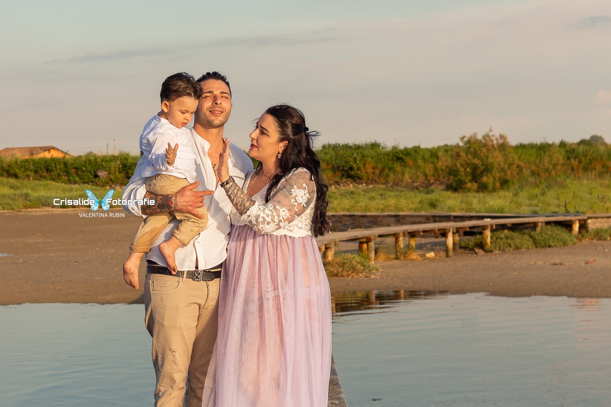 Mamma incinta sulla destra con abito bianco in pizzo e gonna in tulle lilla, mano alzata per dare il cinque al figlio. Papà al centro con camicia bianca e pantaloni beige, in braccio il bambino di circa 1 anno e mezzo con abbigliamento coordinato con papà. Papà sorridente con occhi chiusi illuminato dal sole. Bambino con manina alzata verso la mamma. La famiglia è sul pontile, con mare ai lati, spiaggia e vegetazione sullo sfondo e cielo azzurro