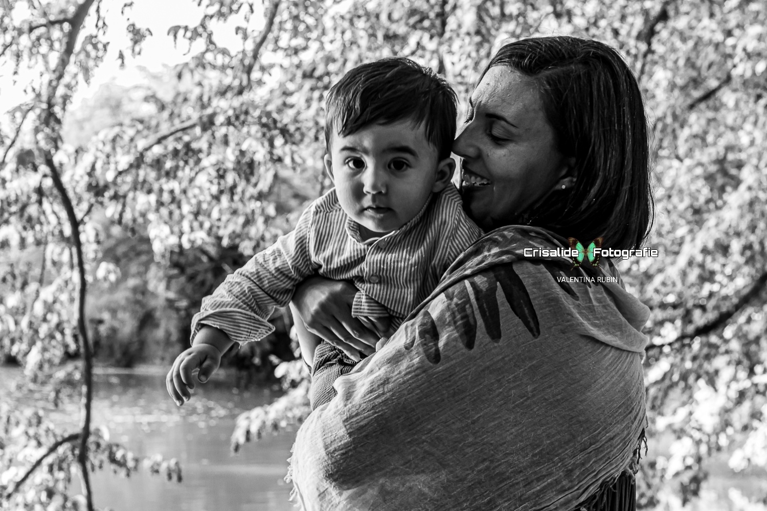 Mamma di profilo con uno scialle sulle spalle sorride guardando il suo bambino in braccio, mentre lui osserva incuriosito verso l'obiettivo, in uno scatto in bianco e nero