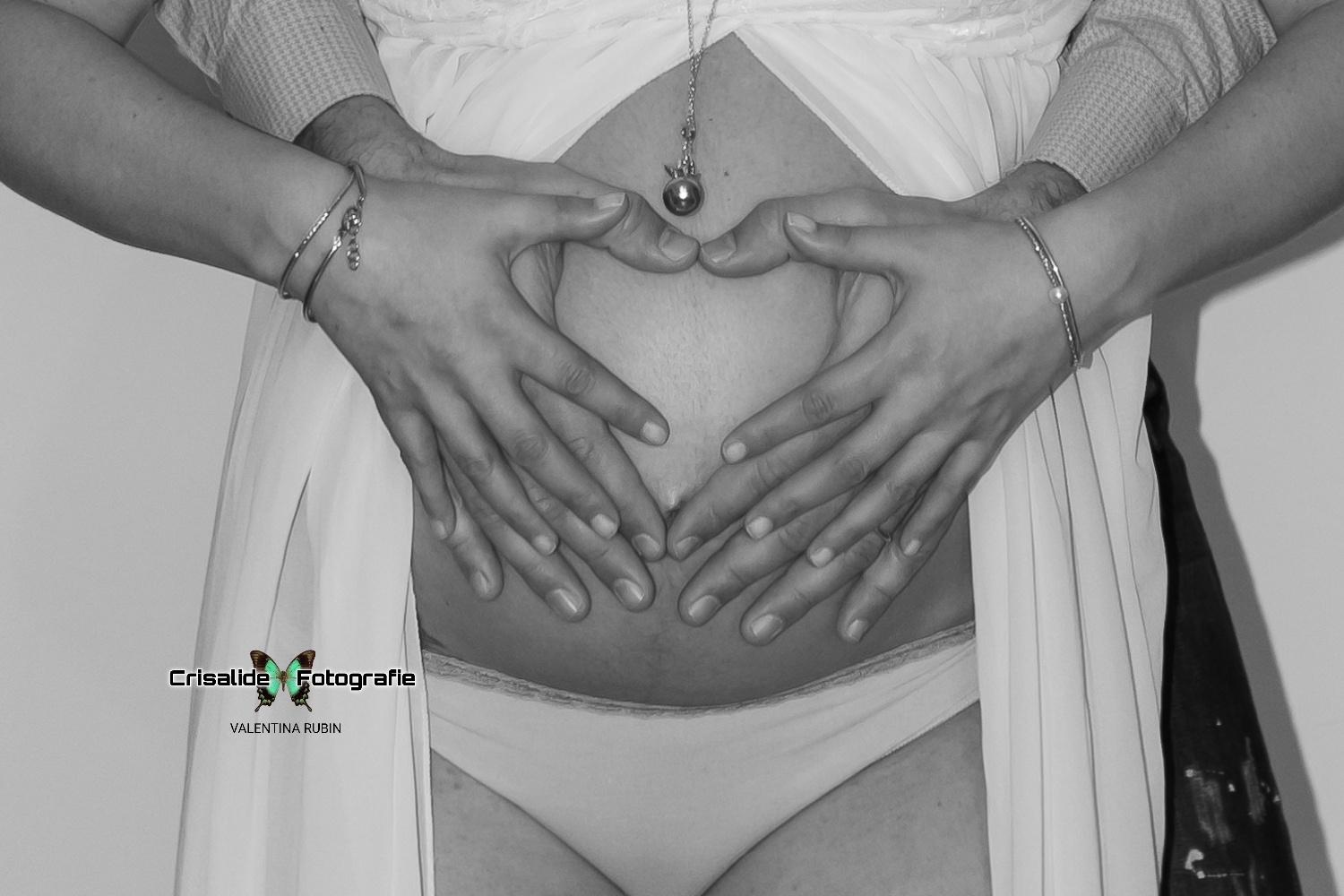 Primo piano del pancione di mamma, mani del papà e della mamma formano un cuore a chiudersi sull'ombelico, ciondolo appena sopra al cuore, braccialetti ai polsi della mamma