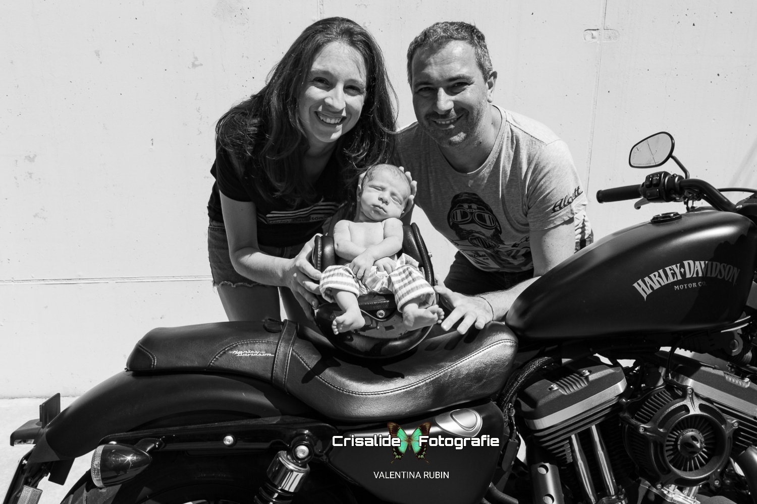 Mamma e papà sorridenti chinati verso il loro neonato che dorme serenamente all'interno di un casco da moto appoggiato su una Harley Davidson. Scatto in bianco e nero realizzato in totale sicurezza, ambientato vicino al garage di casa