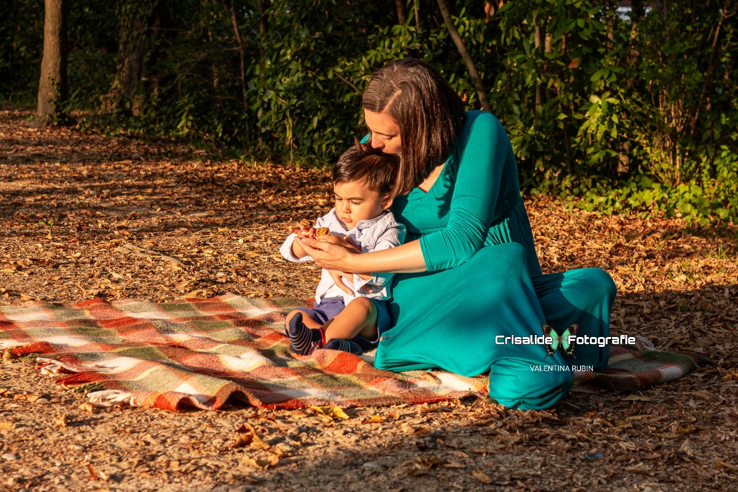 Mamma seduta su una coperta bacia la testa del suo bambino mentre lui tiene una foglia secca, in un ritratto a colori con sfondo autunnale