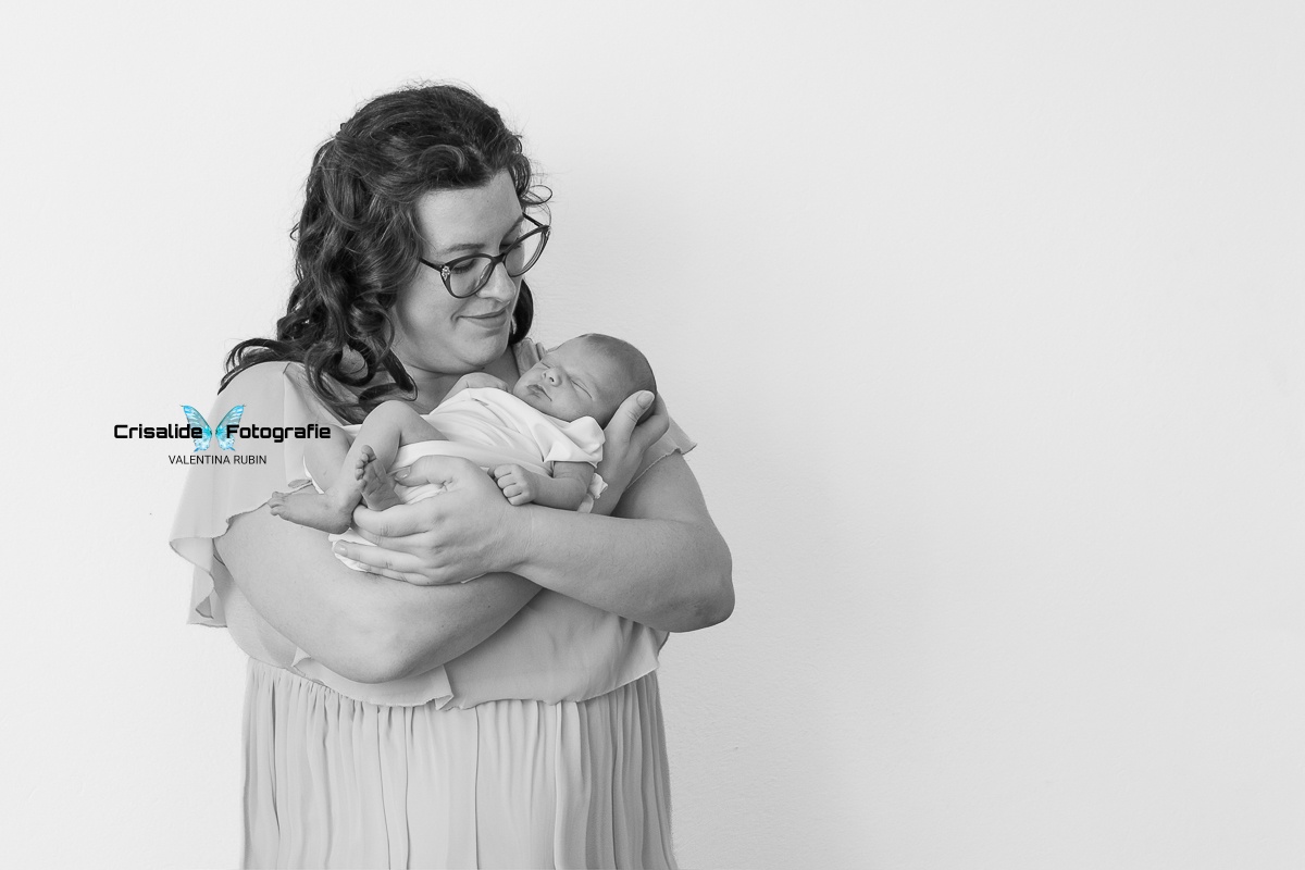 Foto in bianco e nero di mamma con neonata in braccio, mamma sorridente che guarda la bambina. Indossa abito in chiffon rosa a maniche corte, capelli castani ondulati sulle spalle e occhiali. Neonata fasciata in tessuto jersey bianco, dorme tranquilla. Sfondo bianco. Momento dolce e intimo madre-figlia