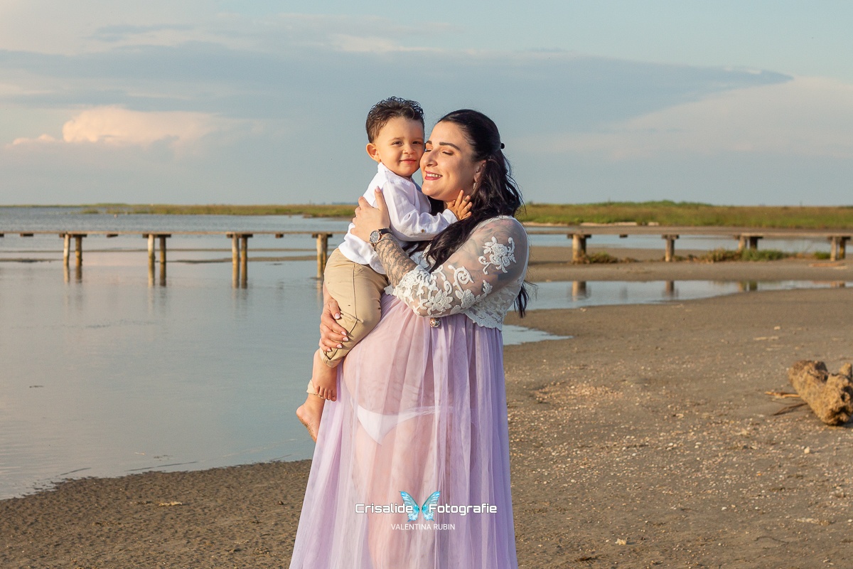 Mamma incinta in abito bianco di pizzo tiene in braccio il bambino vestito di bianco e beige, sorridono, sfondo spiaggia, tronco, mare e pontile