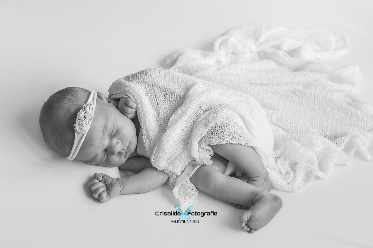 Neonata che dorme avvolta in una tela bianca, con fascetta floreale sulla testa, fotografata in bianco e nero