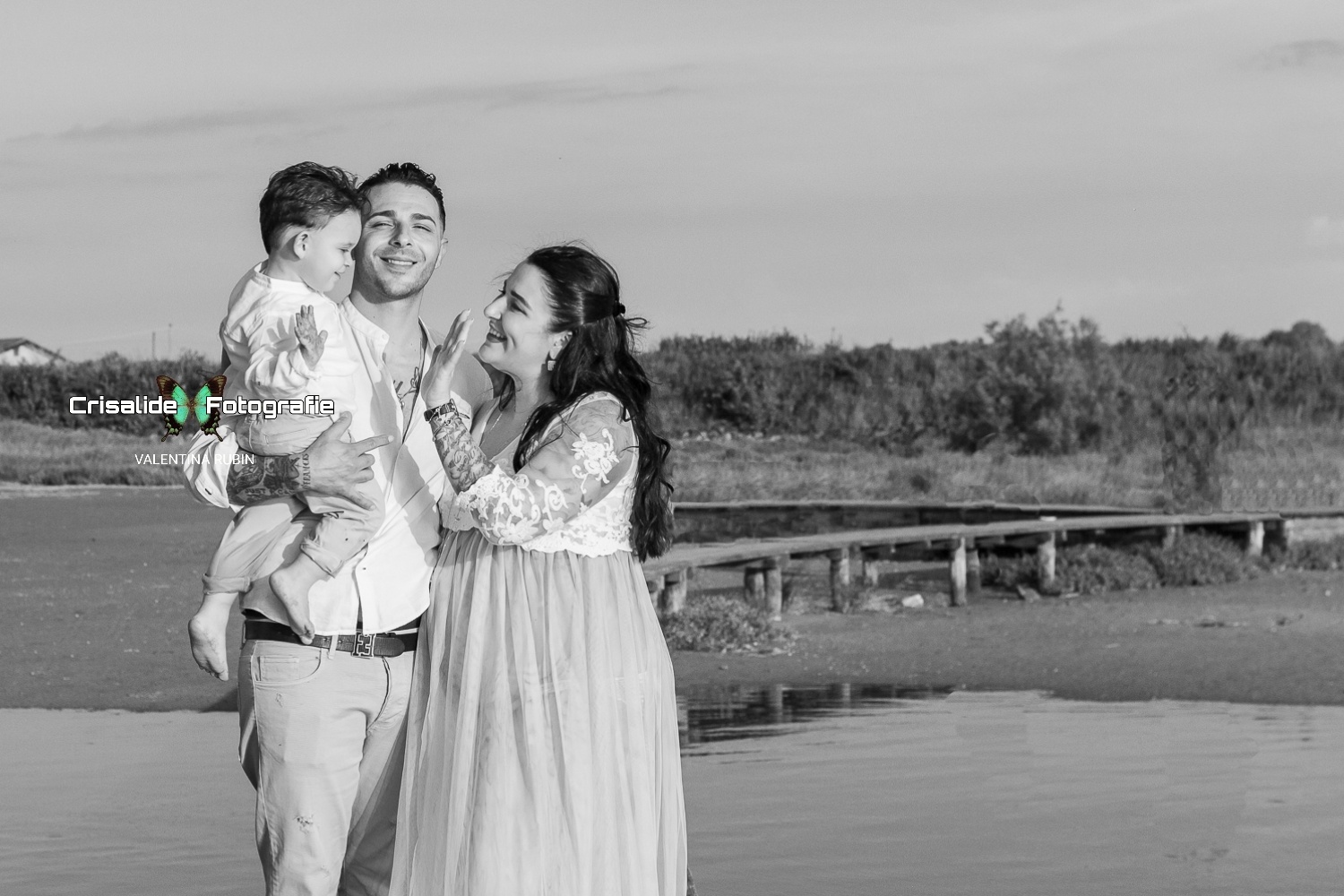 Mamma in abito bianco di pizzo e papà in camicia bianca e pantaloni beige, tiene in braccio il bambino sorridente pronto a dare il cinque alla mamma, sfondo pontile, mare e vegetazione