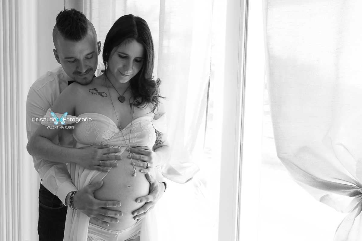 Mamma incinta in chiffon bianco e papà dietro di lei, mani sul pancione, luce entra dalla finestra a sinistra