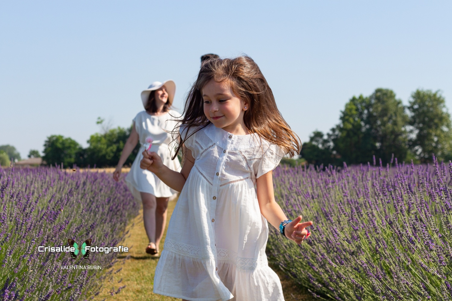 Bambina vestita di bianco in primo piano che guarda felice la lavanda, con i genitori sullo sfondo tra i filari
