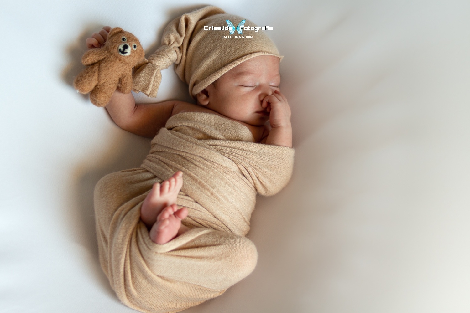 Neonato che dorme a pancia in su, fasciato in beige con berretto coordinato, tiene un orsacchiotto in mano, sfondo bianco