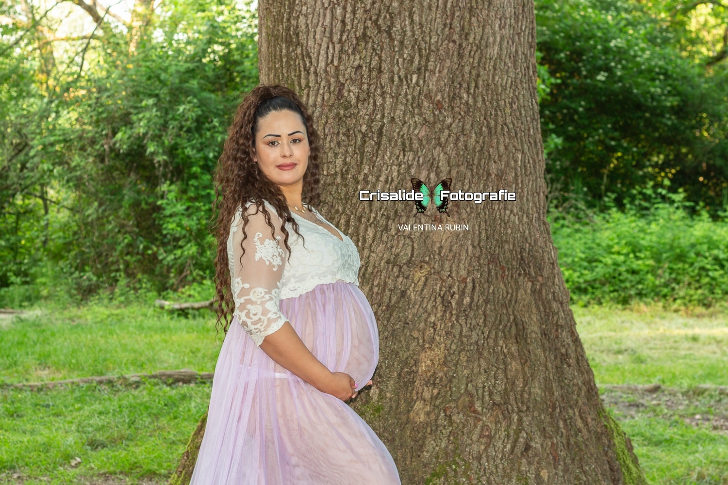 Donna incinta di profilo con vestito bianco in pizzo e gonna in tulle violetto, che sorride all'obiettivo con le mani sotto la pancia, davanti a un tronco di quercia secolare e circondata da alberi e prato verde