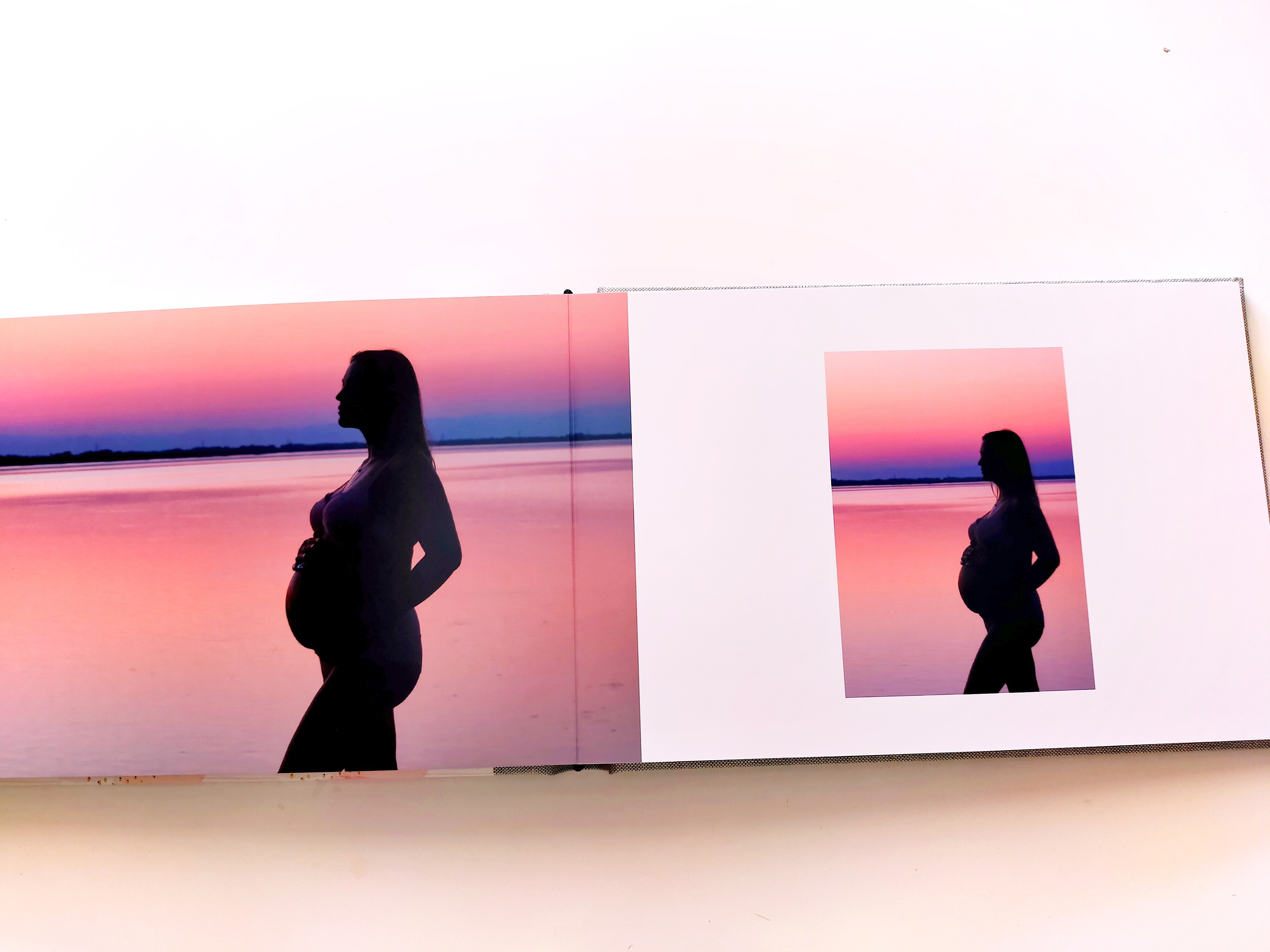 Album fotografico aperto con due foto stampate sulle pagine. Pagina sinistra: foto orizzontale con silhouette di mamma incinta su sfondo mare nei colori rosa, blu e viola. Pagina destra: foto verticale dello stesso soggetto, sfondo mare al tramonto, colori rosa, blu e viola. Sfondo bianco. Visualizzazione del layout interno e qualità di stampa