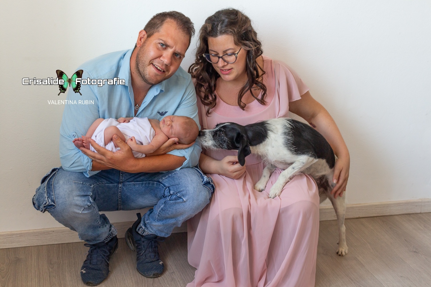 Papà accucciato che tiene in braccio la neonata, mamma accanto con il cane sulle ginocchia che annusa la testina della bambina, momento di famiglia naturale e dolcezza