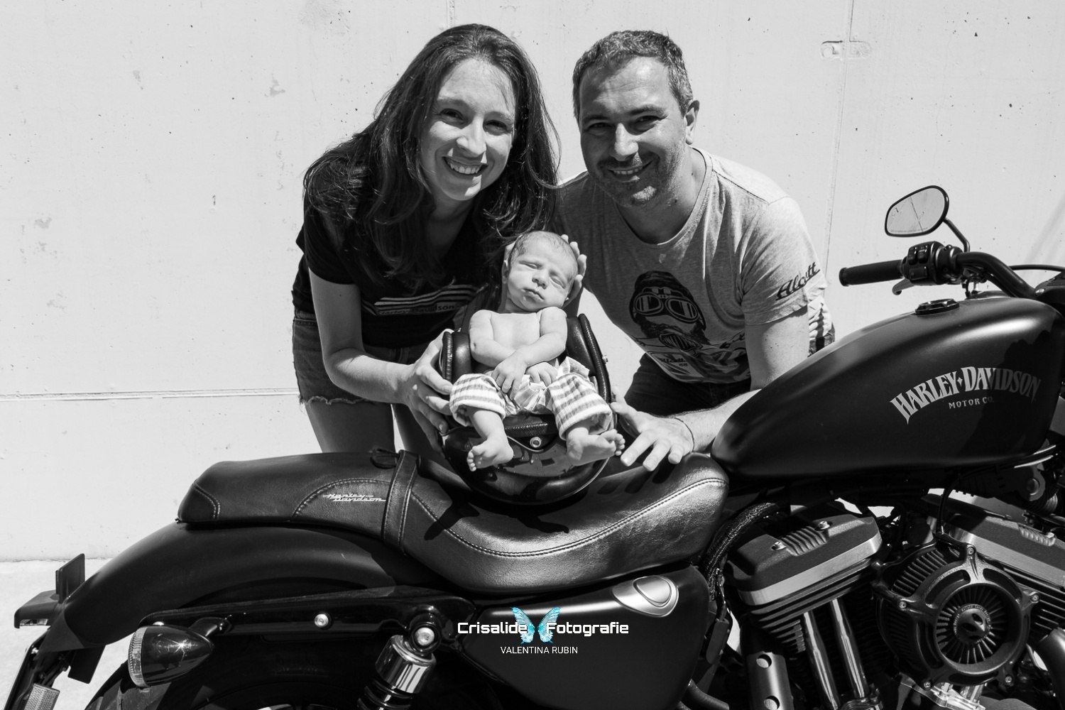 Mamma e papà sorridenti chinati verso il loro neonato che dorme serenamente all'interno di un casco da moto appoggiato su una Harley Davidson. Scatto in bianco e nero realizzato in totale sicurezza, ambientato vicino al garage di casa