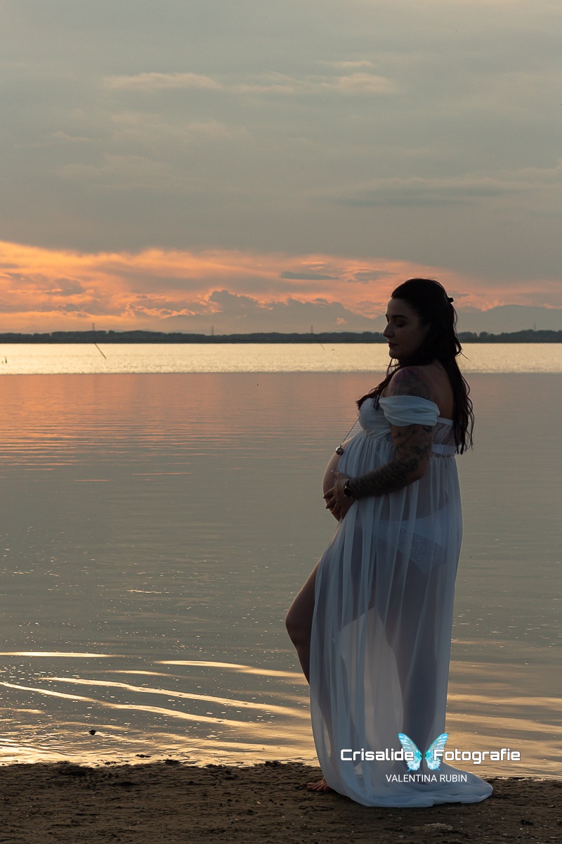 Donna incinta di profilo con abito in chiffon bianco a spalle scoperte, gamba piegata e mano sulla pancia. Capelli neri ondulati sulle spalle, ciondolo sul pancione. Ritratto in penombra sulla spiaggia, piedi sulla sabbia vicino al mare. Sullo sfondo cielo con nuvole grigio-azzurre illuminate da una lieve luce dorata del tramonto