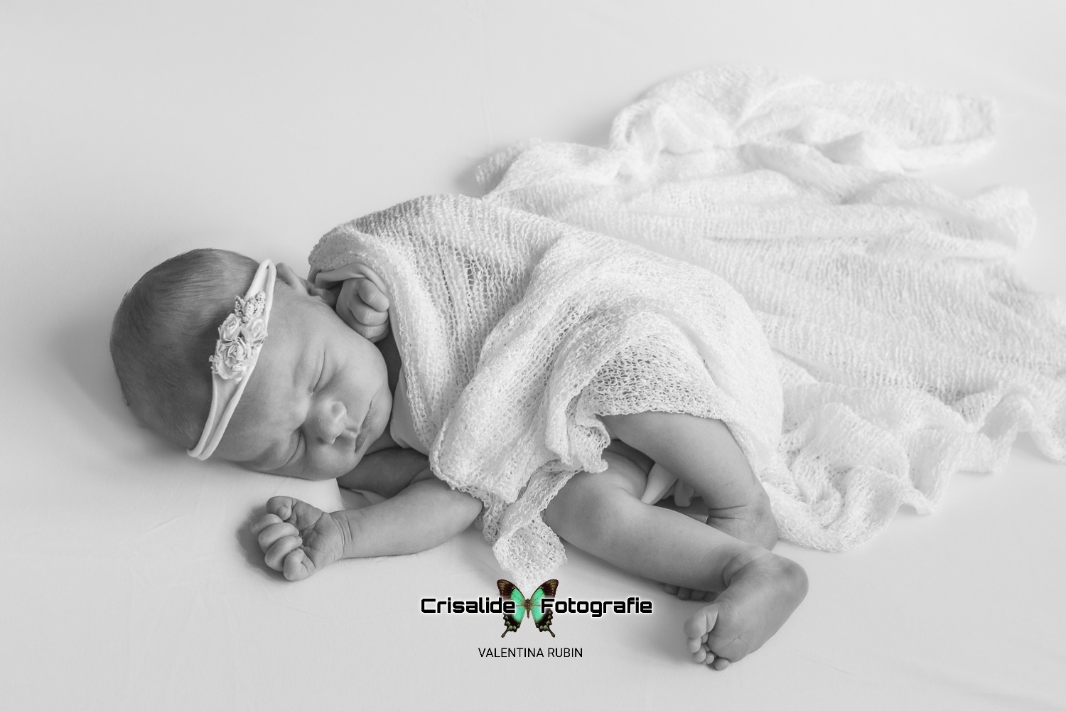 Neonata che dorme avvolta in una tela bianca, con fascetta floreale sulla testa, fotografata in bianco e nero