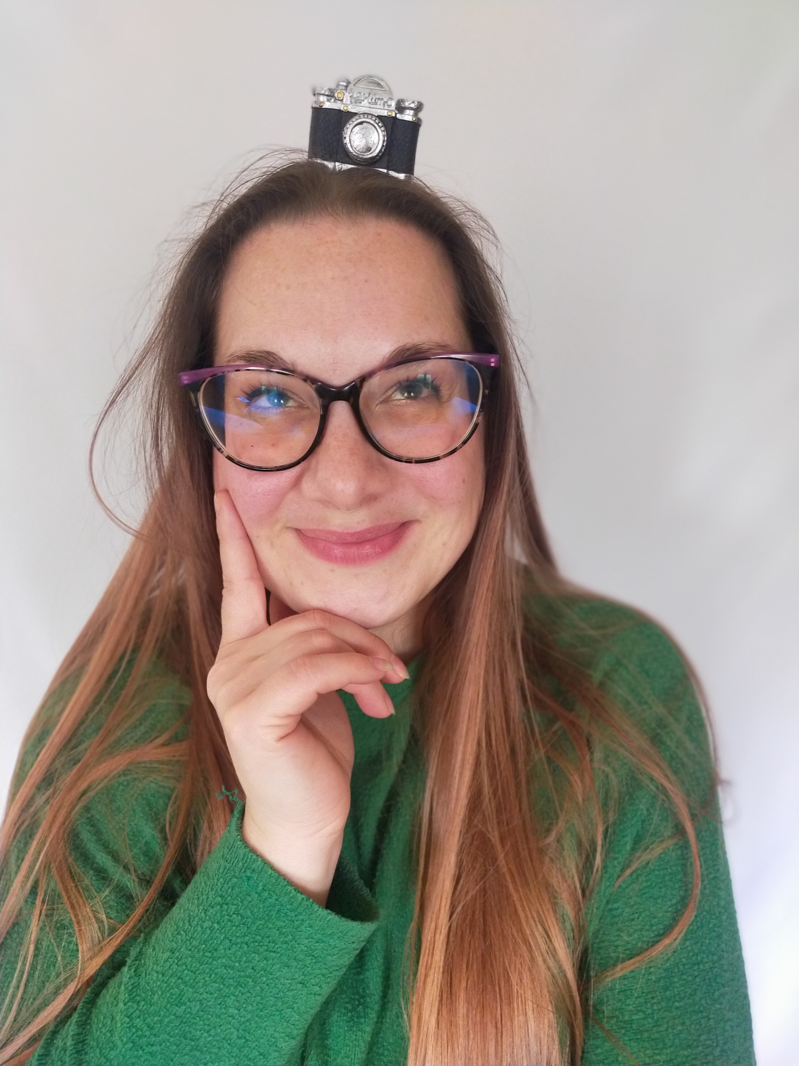 Valentina Rubin, fotografa specializzata in maternità, neonati e famiglie, ritratta con maglione verde, capelli lunghi lisci, occhiali da vista, sulla testa una mini macchina fotografica giocattolo, sguardo in alto verso la fotocamera sulla testa, sorriso luminoso, sfondo bianco