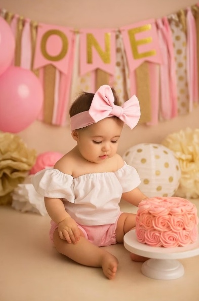 Bambina seduta a terra, con pantaloncini rosa, magliettina bianca, fiocco rosa in testa, guarda la torta che ha davanti. sfondo colorato