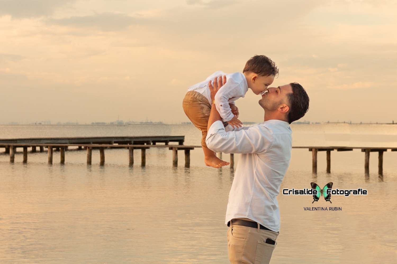 Papà in camicia bianca e pantaloni beige tiene in braccio il bambino vestito uguale, mentre lo bacia, sfondo pontile e mare con nuvole