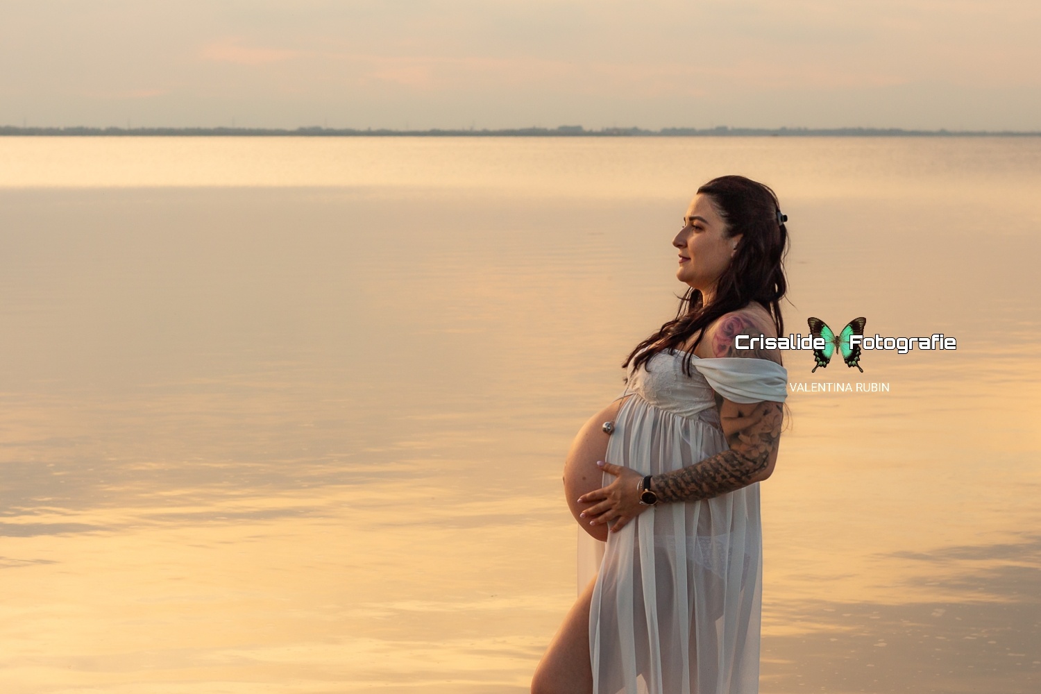 Mamma incinta di profilo in abito di chiffon bianco aperto sul pancione, con il mare sullo sfondo e riflessi rosa pastello sull'acqua