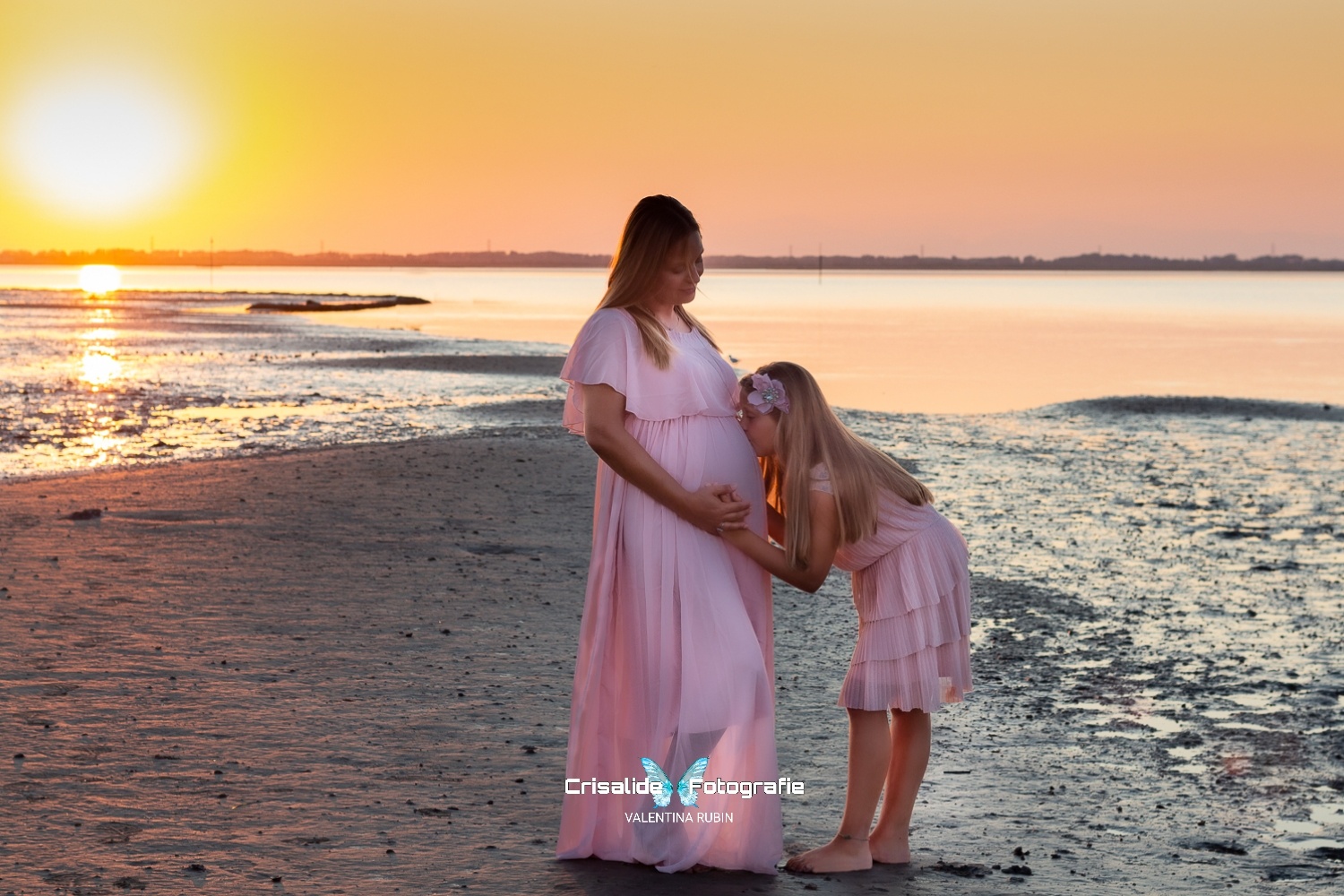 Mamma incinta in abito rosa sulla spiaggia con la figlia di 8 anni, anche lei vestita di rosa. La bambina bacia la pancia della mamma e appoggia una mano sopra, mentre la mamma sorride e poggia la sua mano sopra quella della figlia. Capelli lisci della mamma sulle spalle, capelli biondi della bambina con fiore tra i capelli. Sfondo mare con riflessi dorati del sole all'orizzonte