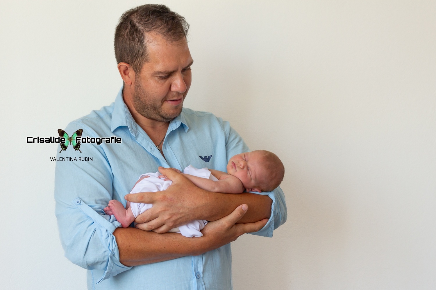 Papà in camicia azzurra che guarda la neonata addormentata tra le sue braccia, su sfondo bianco