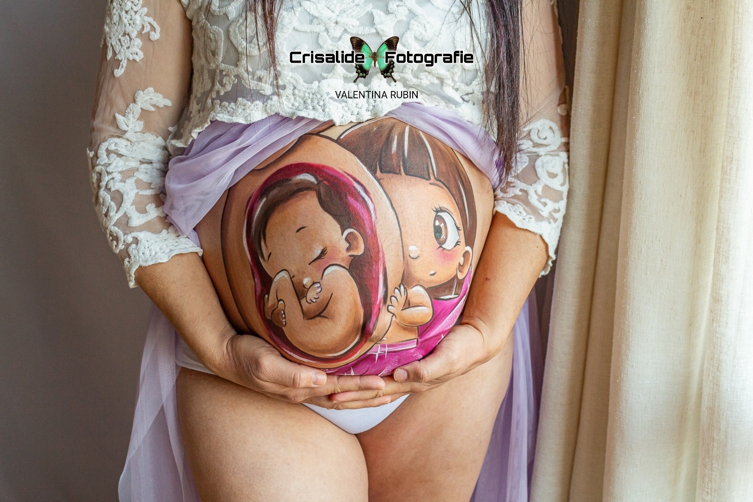 Primo piano sul pancione di una mamma con belly painting realizzato a mano da un'artista. Mamma con abito bianco in pizzo, manica tre quarti, gonna in tulle viola aperta sulla pancia. Entrambe le mani sotto il ventre, dita intrecciate. Luce morbida laterale proveniente dalla finestra, tenda sullo sfondo, atmosfera intima e delicata