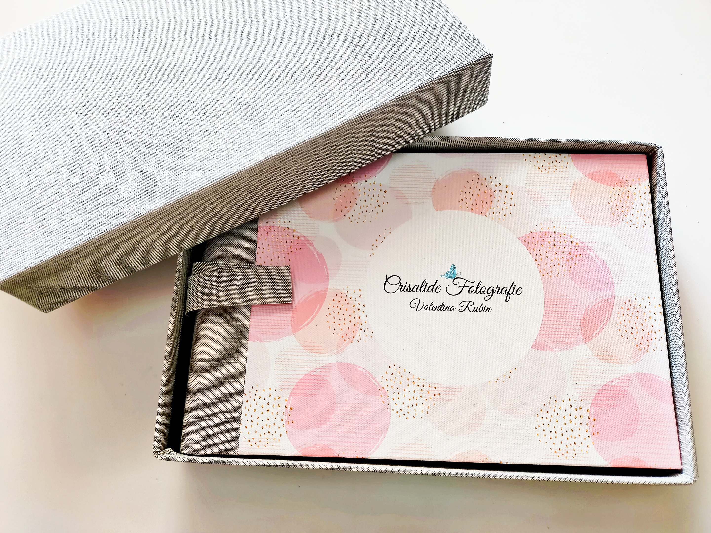 Album fotografico con copertina rigida rosa con logo, posizionato dentro un box di cartone rivestito in tessuto grigio. Coperchio del box in cartone grigio visibile nell'angolo superiore sinistro. Sfondo bianco. Presentazione elegante e professionale del prodotto