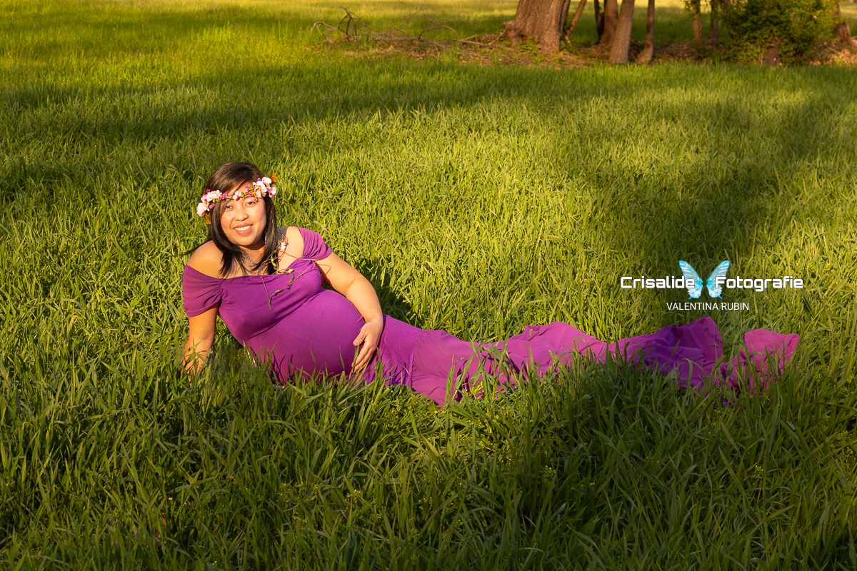 Donna filippina incinta sdraiata sul fianco destro, sull'erba, con abito viola a sirena e spalle scoperte, capelli neri lunghi, sorridente e guardando in camera; mano destra sull'erba e l'altra sulla pancia; illuminata da luce dorata del tramonto con ombre degli alberi; sfondo sfocato con accenno di alberi