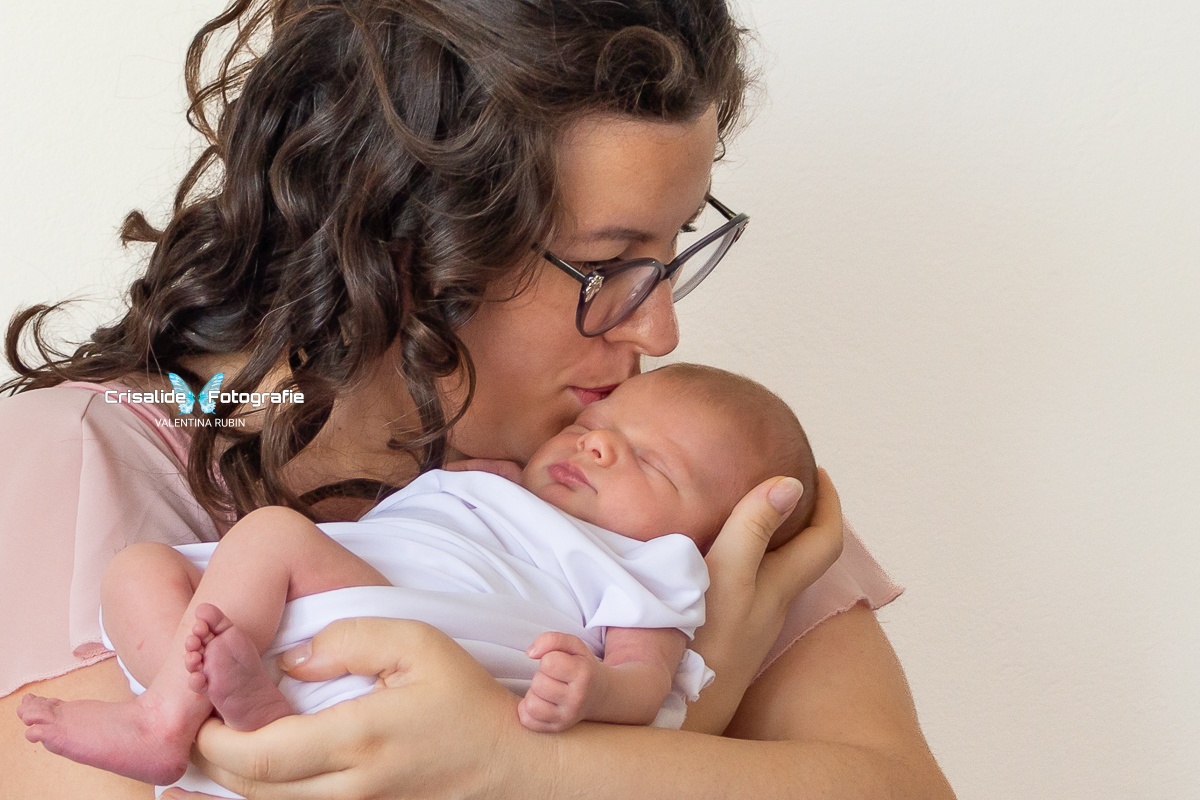 Mamma con capelli castani ondulati e occhiali che bacia la testa della sua neonata addormentata, avvolta in una fascia bianca, su sfondo bianco
