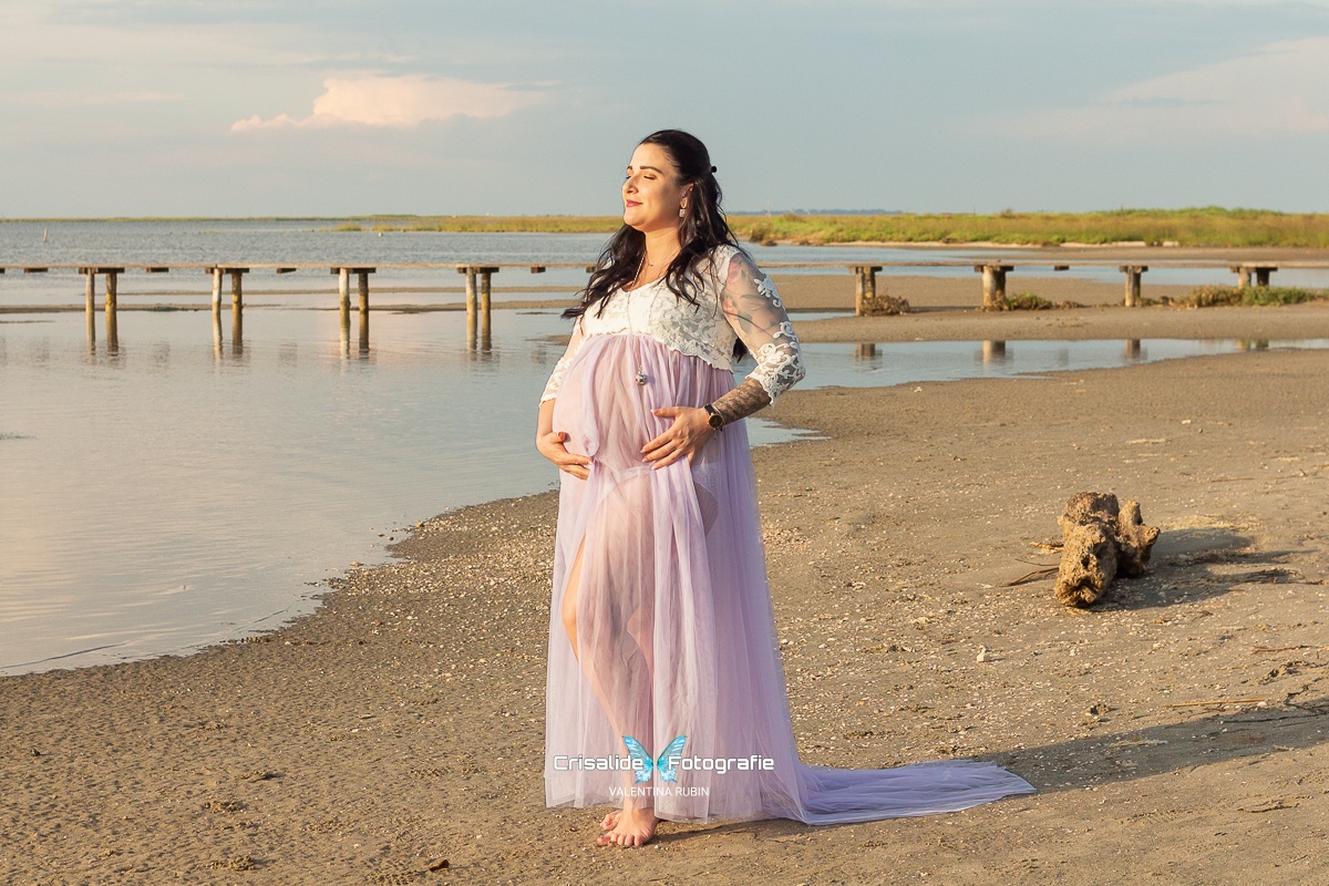 Mamma incinta con abito bianco in pizzo e gonna in tulle violetto, fotografata di tre quarti mentre tiene le mani sulla pancia. Ha gli occhi chiusi rivolti verso il sole. Sfondo con mare a sinistra, spiaggia con un tronco a destra, pontile di legno in lontananza ed erba. Cielo azzurro con una nuvola bianca