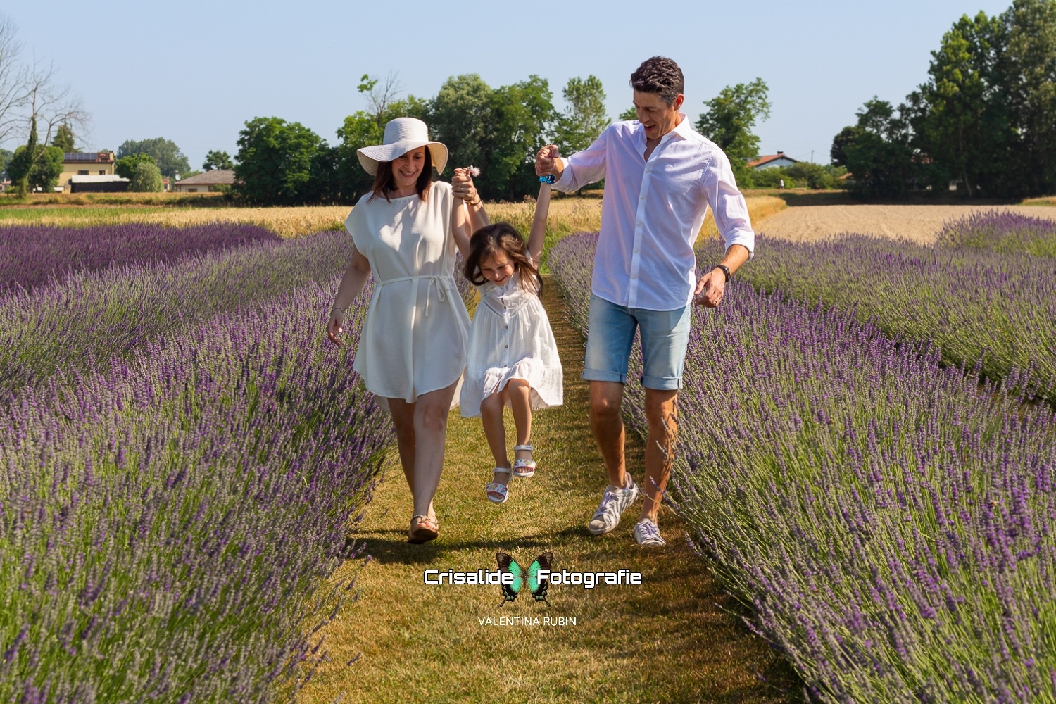 Mamma e papà vestiti di bianco sollevano la bambina per mano facendola saltare tra i filari di lavanda, tutti sorridenti