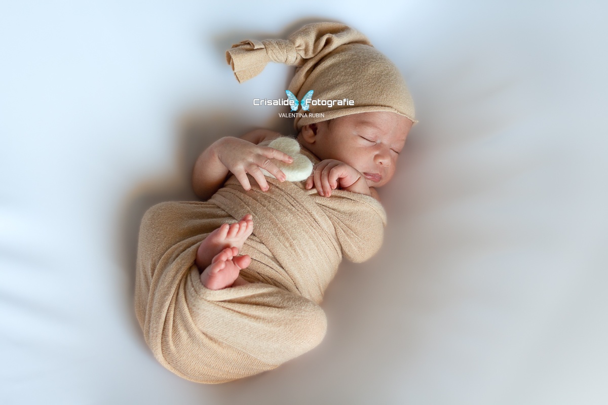Neonato che dorme supino avvolto in fascia di cotone beige, con gambine rannicchiate, manina sulla guancia, cuore bianco in feltro nella mano, berrettino coordinato e sfondo bianco - fotografia newborn professionale a domicilio