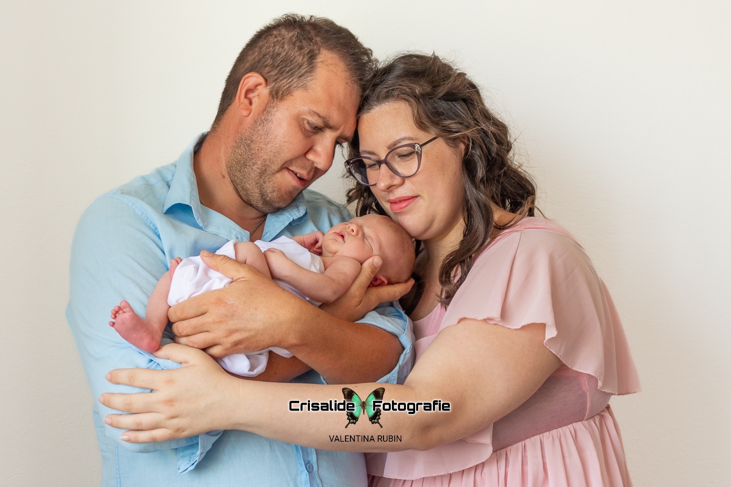 Papà con camicia azzurra tiene in braccio una neonata fasciata di bianco che dorme, mamma con abito in chiffon rosa, maniche corte, occhiali e capelli marroni con boccoli accarezza la bambina poggiando il mento sulla sua testa; mani dei genitori vicine, teste che si toccano; sfondo bianco con luce morbida da finestra laterale, catturando sorrisi e gesti spontanei in stile fotografico autentico