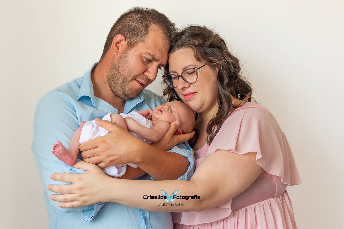 Papà con camicia azzurra tiene in braccio una neonata fasciata di bianco che dorme, mamma con abito in chiffon rosa, maniche corte, occhiali e capelli marroni con boccoli accarezza la bambina poggiando il mento sulla sua testa; mani dei genitori vicine, teste che si toccano; sfondo bianco con luce morbida da finestra laterale, catturando sorrisi e gesti spontanei in stile fotografico autentico