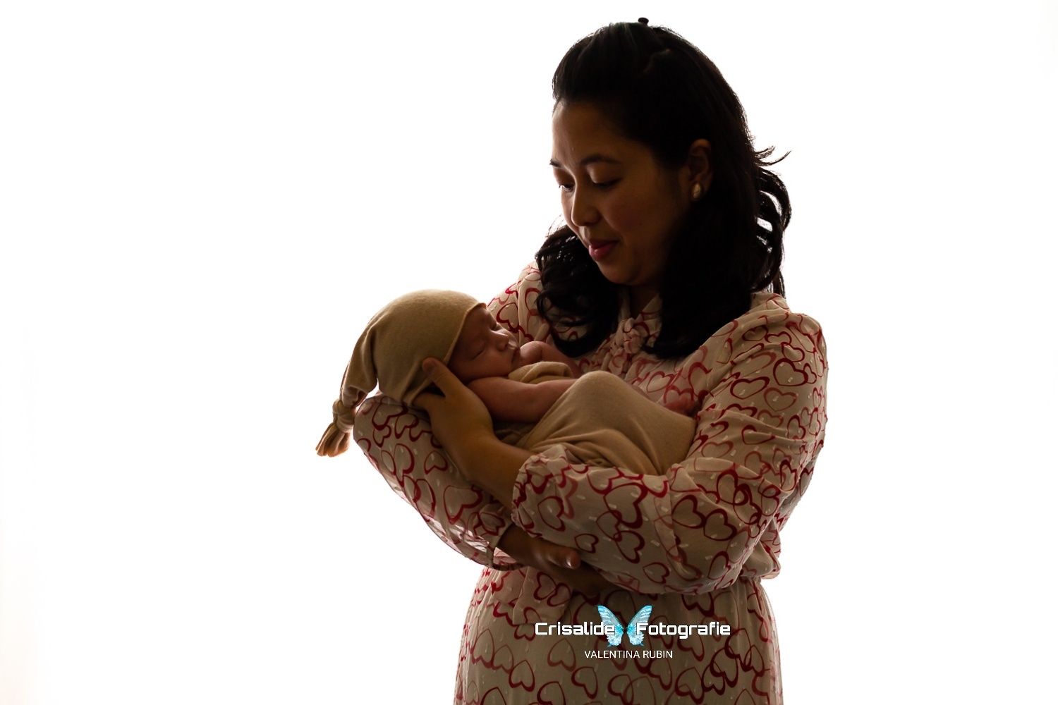 Mamma in abito beige con cuori rossi tiene in braccio il neonato fasciato in beige con berretto coordinato, sfondo bianco