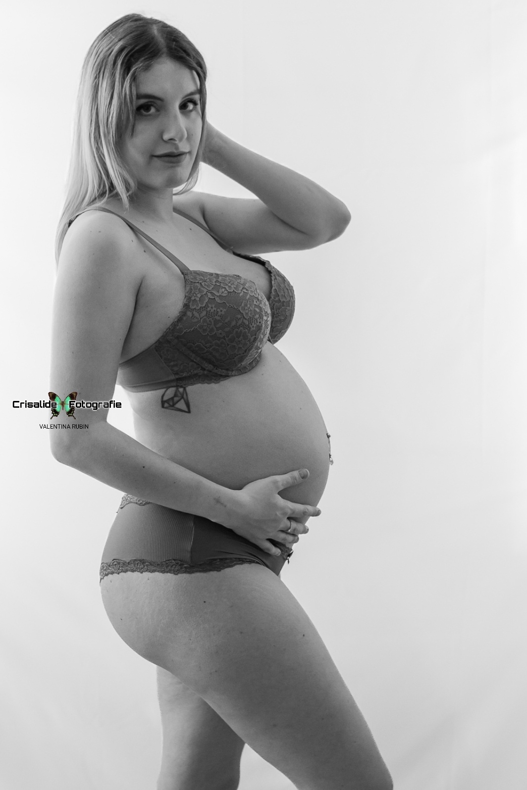 Mamma incinta di profilo in intimo scuro, gamba piegata, una mano sotto la pancia, e una dietro la testa, sguardo in camera, luce laterale su sfondo bianco