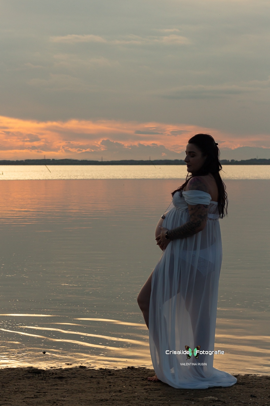 Donna incinta di profilo con abito in chiffon bianco a spalle scoperte, gamba piegata e mano sulla pancia, fotografata alla spiaggia della Boschettona a Codevigo, in provincia di Padova. Capelli neri ondulati sulle spalle, ciondolo sul pancione. Ritratto in penombra sulla spiaggia, piedi sulla sabbia vicino al mare. Sullo sfondo cielo con nuvole grigio-azzurre illuminate da una lieve luce dorata del tramonto