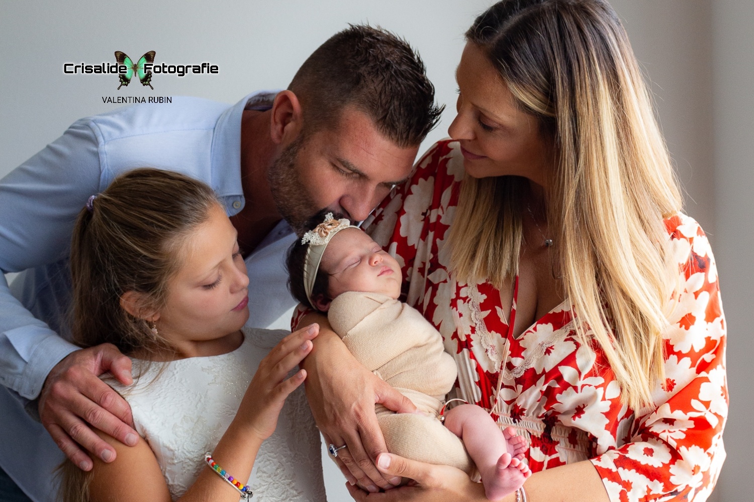 Ritratto di famiglia, con mamma, papà, neonata e sorellina, La neonata è al centro della scena, in braccio alla mamma che è sulla destra, guarda il papà sorridente. Il papà dietro bacia la testina della neonata. La sorellina sulla sinistra tocca la mano della mamma vicino alla neonata. Atmosfera accogliente, richiama casa