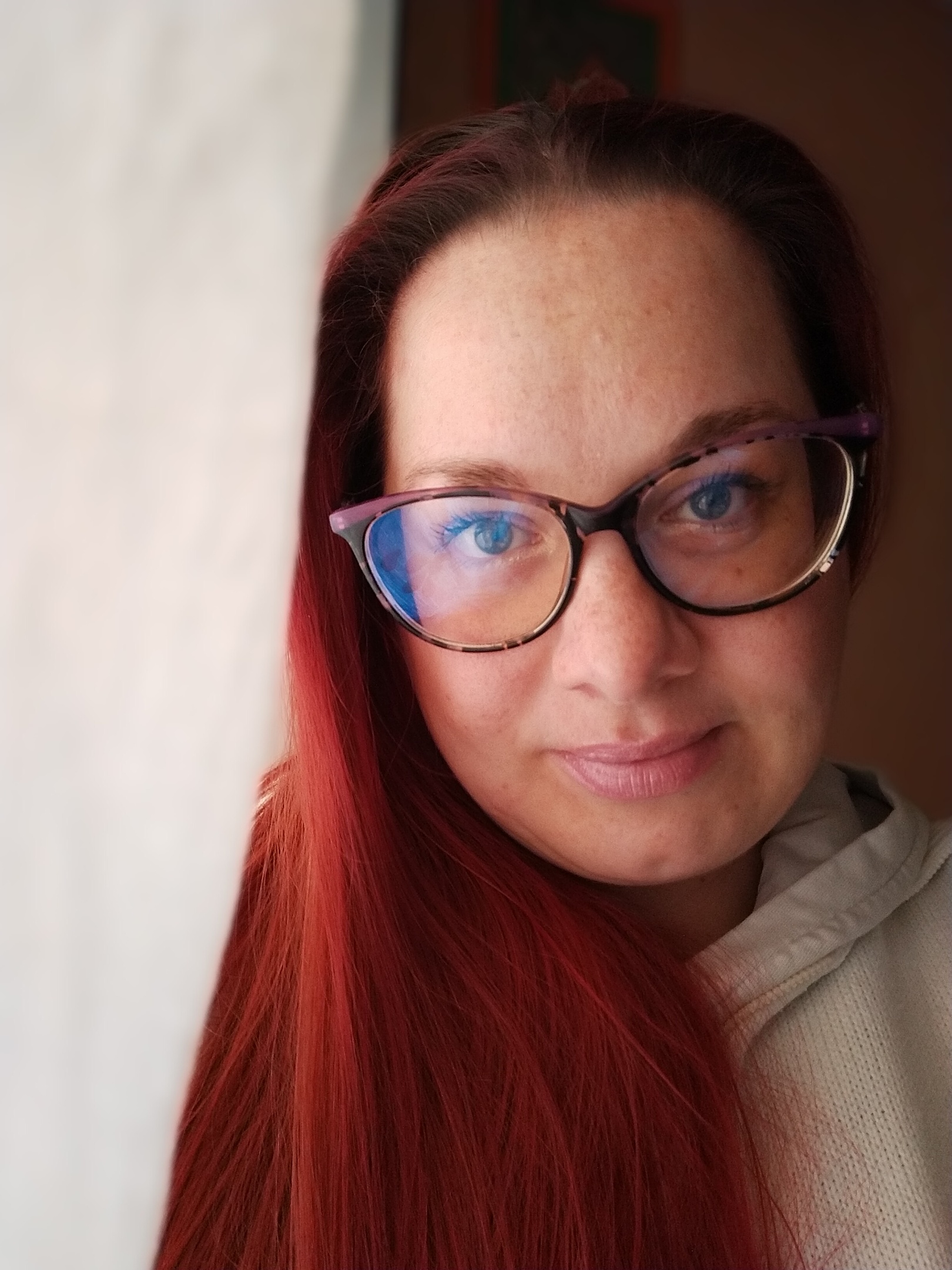 Valentina Rubin, fotografa specializzata in maternità, neonati e famiglie, ritratta con capelli rossi lisci, occhiali e un leggero sorriso, in luce calda e morbida