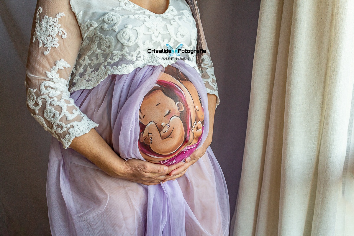 Donna incinta con vestito bianco in pizzo e gonna il tulle violetto, che accarezza la pancia con belly painting, illuminata dalla finestra di casa