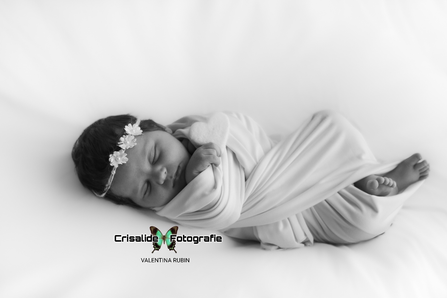 Neonata che dorme fasciata di bianco, a figura intera, con cuore bianco in mano e coroncina di fiori, scatto in bianco e nero