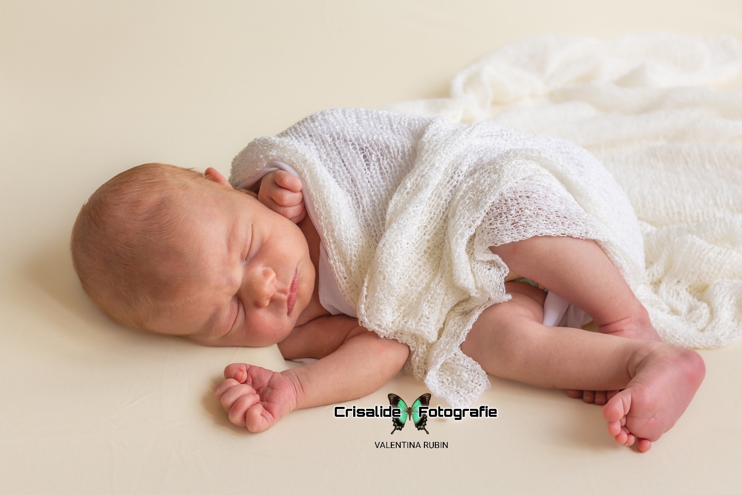 Neonata che dorme di lato, avvolta in tela bianca, con pugnetti e gambine visibili, sfondo color panna - fotografia newborn professionale a domicilio, servizio Pacchetto Mini