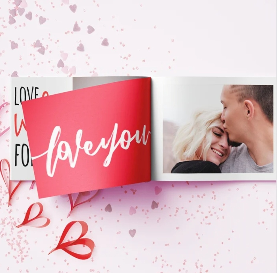 Fotolibro aperto con scritta love you su sfondo rosso e foto stampata di una coppia, decorazioni a forma di cuore sullo sfondo