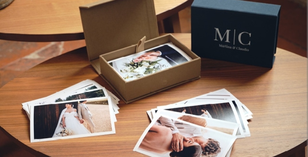 Cofanetto in cartone con stampe fotografiche all'interno e sparse sul tavolo. Coperchio appoggiato sul tavolo con logo personalizzato. Contenuto visibile: foto in alta qualità, scatola elegante e ordinata
