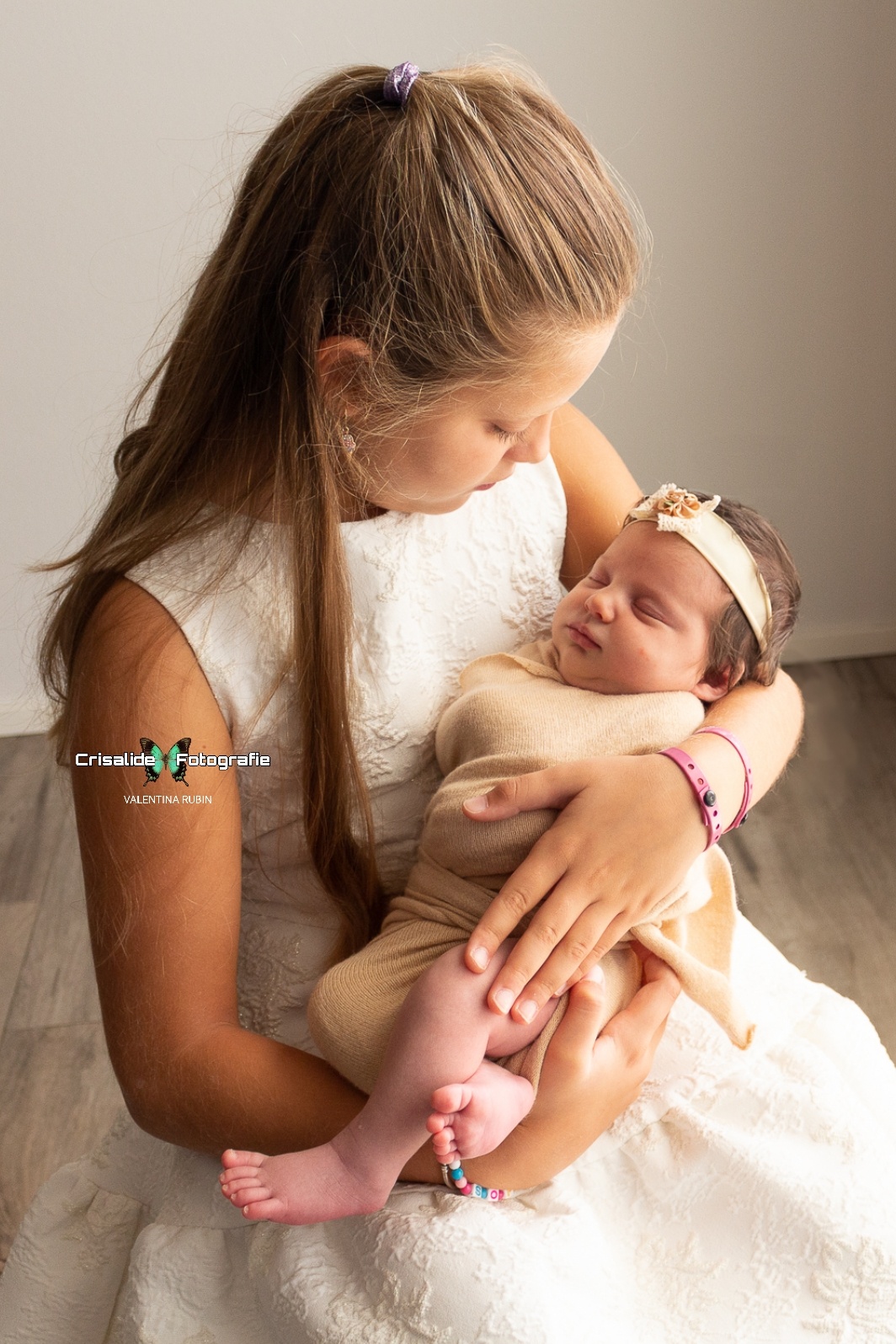 Sorella maggiore vestita di bianco che tiene in braccio la neonata fasciata di beige mentre la guarda dormire, su sfondo chiaro