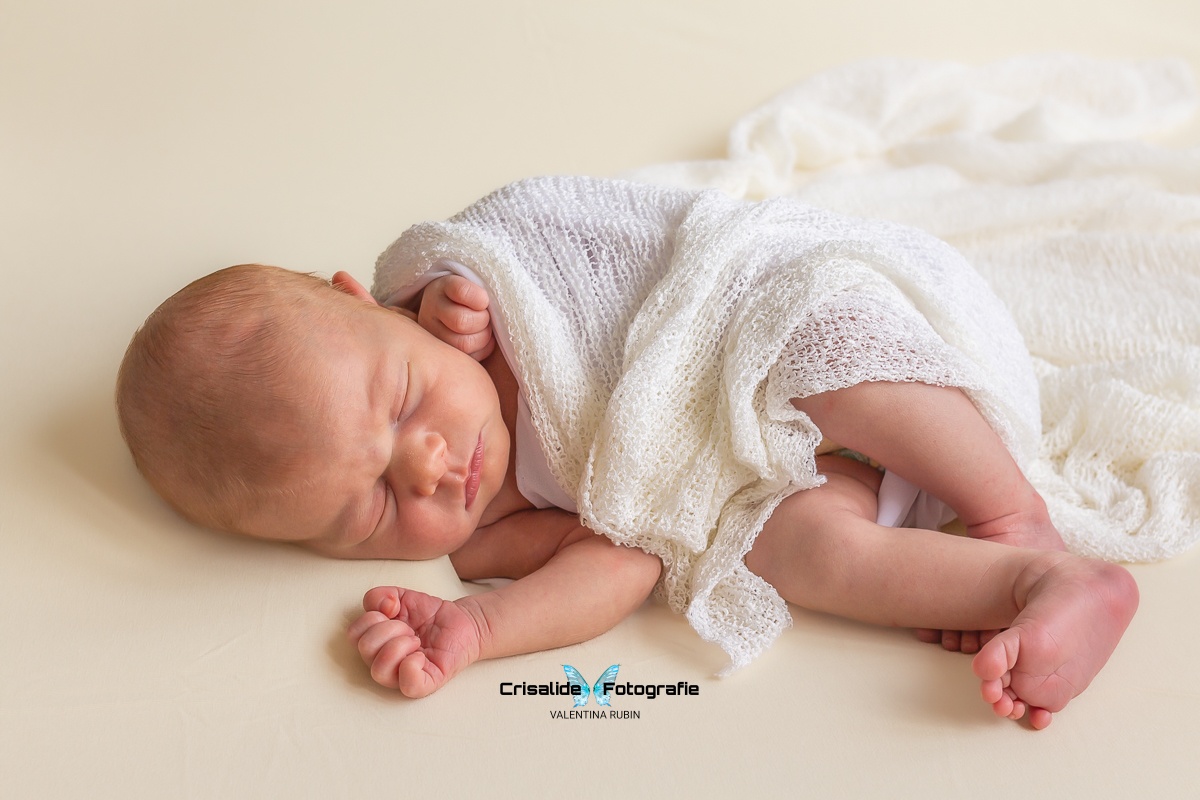 Neonata che dorme di lato, avvolta in tela bianca, con pugnetti e gambine visibili, sfondo color panna - fotografia newborn professionale a domicilio, servizio Pacchetto Mini