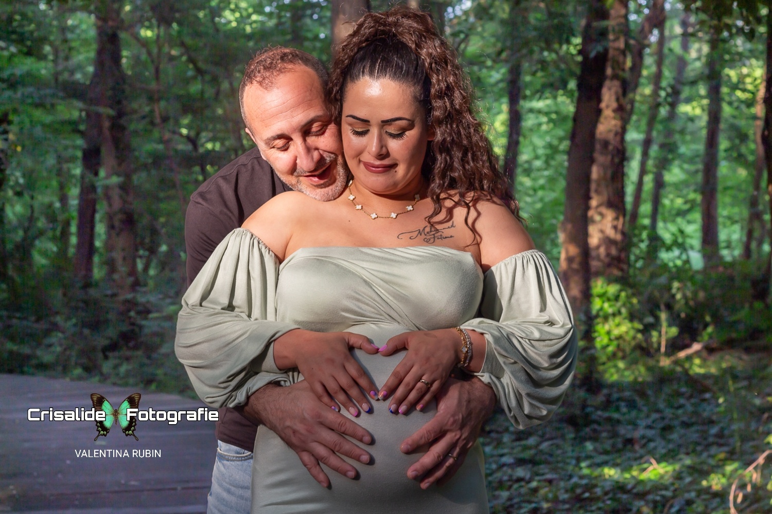 Mamma incinta, con marito che l'abbraccia da dietro, lui tiene la testa sulla spalla di lei, entrambi con le mani e sguardo sul pancione. Ambiente esterno in mezzo alla natura alberi e verde