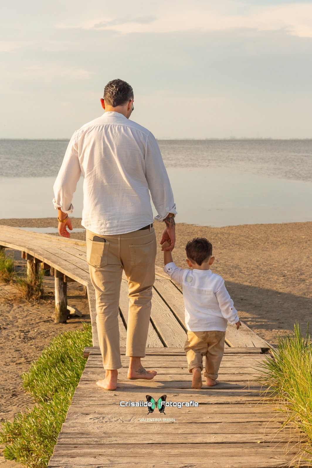 Papà e figlio che si tengono per mano mentre camminano, di spalle dall'obiettivo, sono sopra un pontile di legno e vanno verso il mare. Sfondo naturale spiaggia e mare, con luce dorata del tramonto