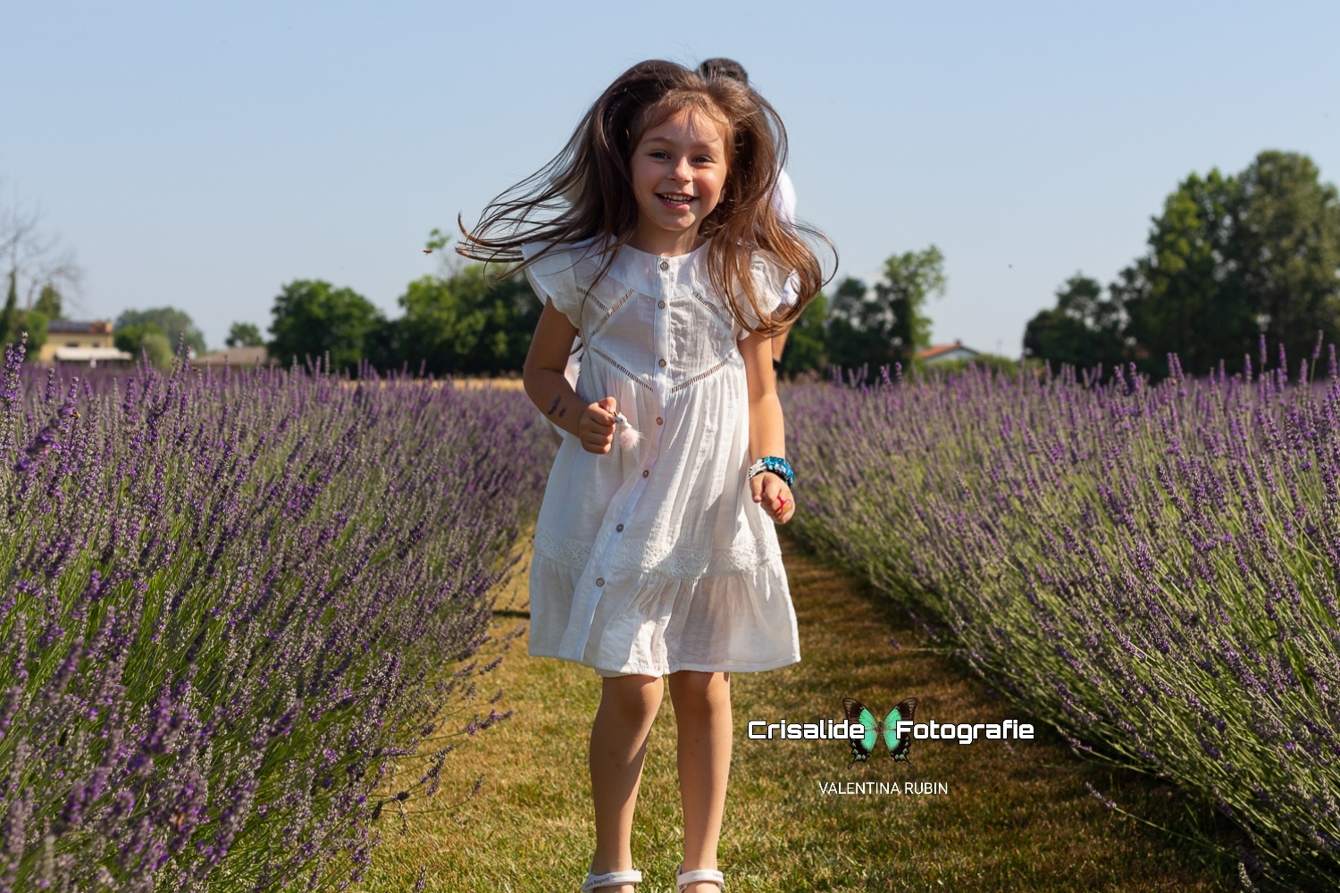 Bambina che salta sorridente tra i filari di lavanda con vestito bianco e capelli che svolazzano