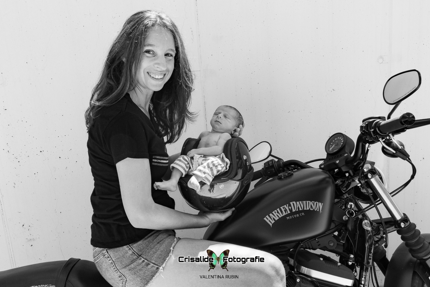 Mamma sorridente seduta sulla sua moto Harley Davidson che guarda in camera mentre tiene il casco con il suo bambino seduto all'interno, in un ritratto in bianco e nero su sfondo bianco