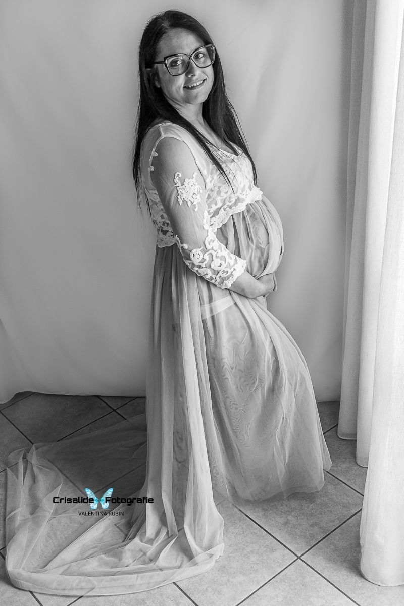 Mamma incinta di profilo in abito bianco di pizzo e gonna lilla in tulle, sguardo in camera e sorriso, occhiale, luce dalla finestra sulla tenda